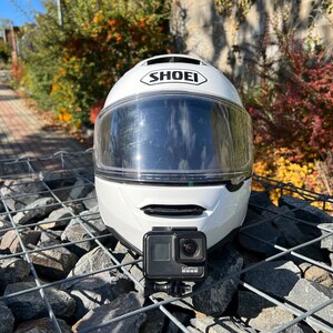 May include: White motorcycle helmet with a clear visor and a GoPro camera attached to the top. The helmet is sitting on a pile of rocks.