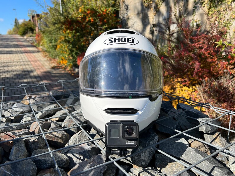 May include: White motorcycle helmet with a clear visor and a GoPro camera attached to the top. The helmet is sitting on a pile of rocks.