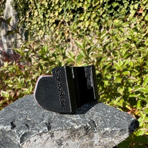 May include: Black 3D printed phone holder with the text "City Pic Rider" on the side. The holder is sitting on a gray rock.