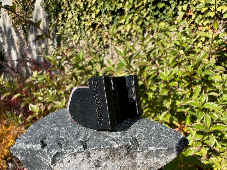 May include: Black 3D printed phone holder with the text "City Pic Rider" on the side. The holder is sitting on a gray rock.