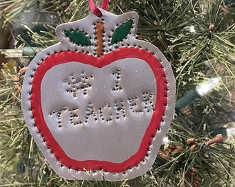 May include: A silver apple-shaped Christmas bauble with a red outline and green leaves. The bauble has the words "#1 Teacher" written on it.