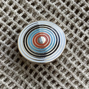Può includere: Uno spillo rotondo dorato con cerchi concentrici di colore nero, bianco, blu, arancione e rosso.