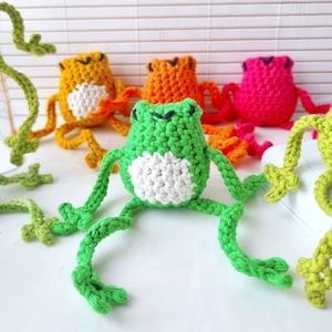 May include: Four crocheted frog toys in different colors: green, orange, yellow, and pink. The green frog is in the foreground and has white eyes and a white belly.