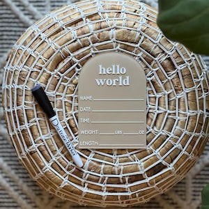 May include: A beige acrylic birth announcement sign with the words "hello world" in white. The sign includes spaces to write the baby's name, date, time, weight (lbs, oz), and length. A black and white dry erase marker is next to the sign.