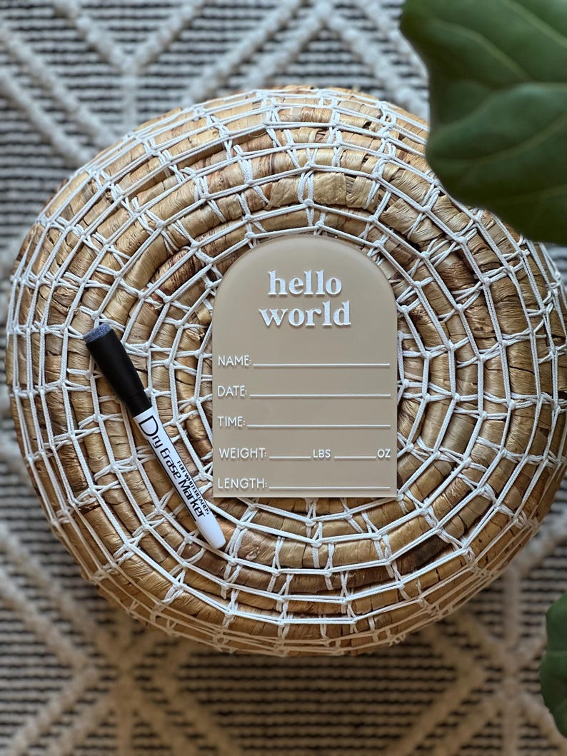 May include: A beige acrylic birth announcement sign with the words "hello world" in white. The sign includes spaces to write the baby's name, date, time, weight (lbs, oz), and length. A black and white dry erase marker is next to the sign.