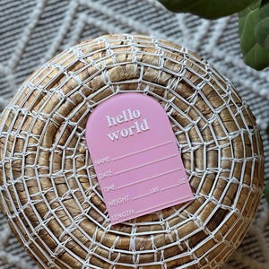 May include: A pink baby announcement tag with the words "hello world" in white. The tag includes spaces to write the baby's name, date, time, weight (lbs), and length (oz). The tag is placed on a woven basket.