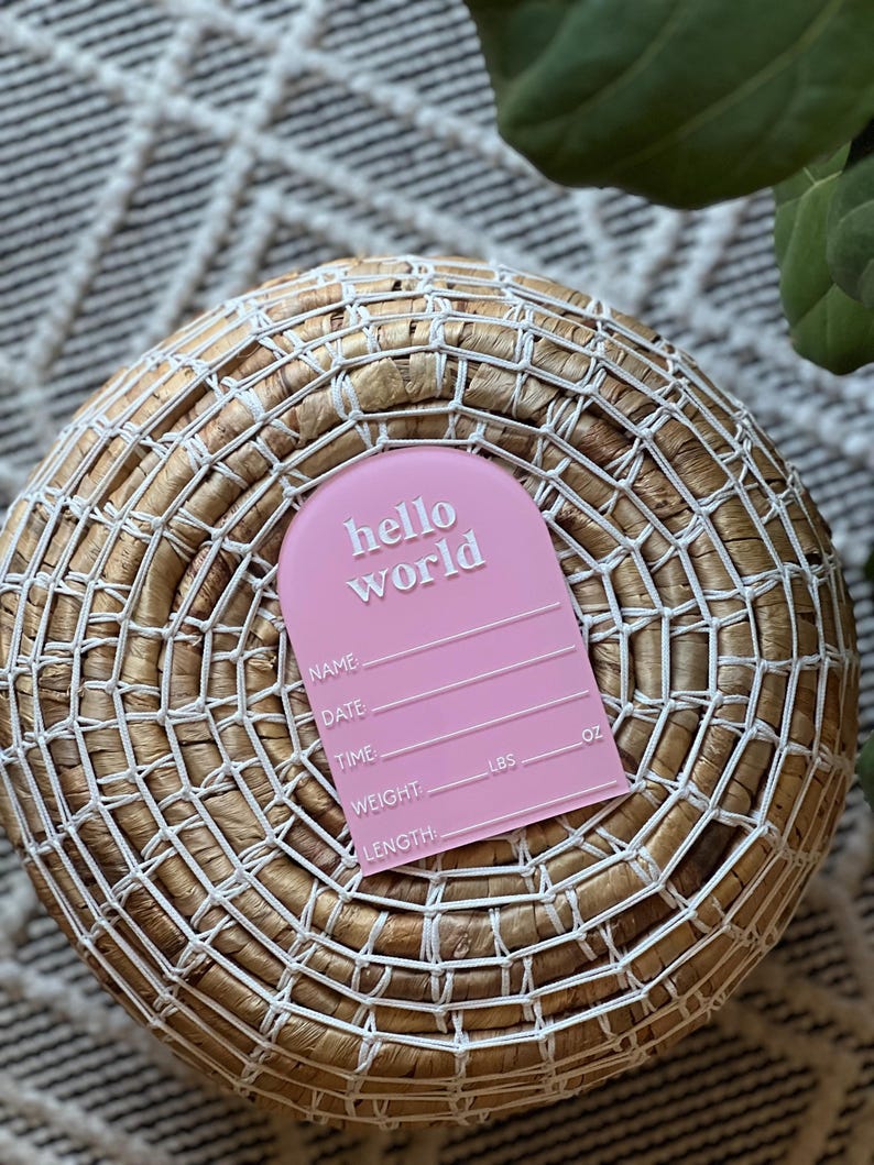May include: A pink baby announcement tag with the words "hello world" in white. The tag includes spaces to write the baby's name, date, time, weight (lbs), and length (oz). The tag is placed on a woven basket.