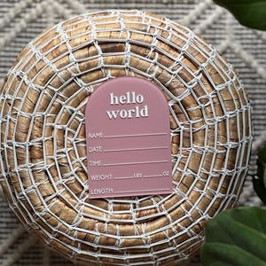 May include: A dusty rose-colored birth announcement plaque with the words "hello world" in white script. The plaque includes spaces to record the baby's name, date, time, weight (lbs, oz), and length. The plaque is placed on a woven basket.
