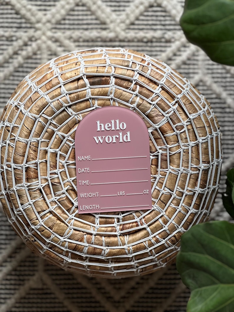 May include: A dusty rose-colored birth announcement plaque with the words "hello world" in white script. The plaque includes spaces to record the baby's name, date, time, weight (lbs, oz), and length. The plaque is placed on a woven basket.