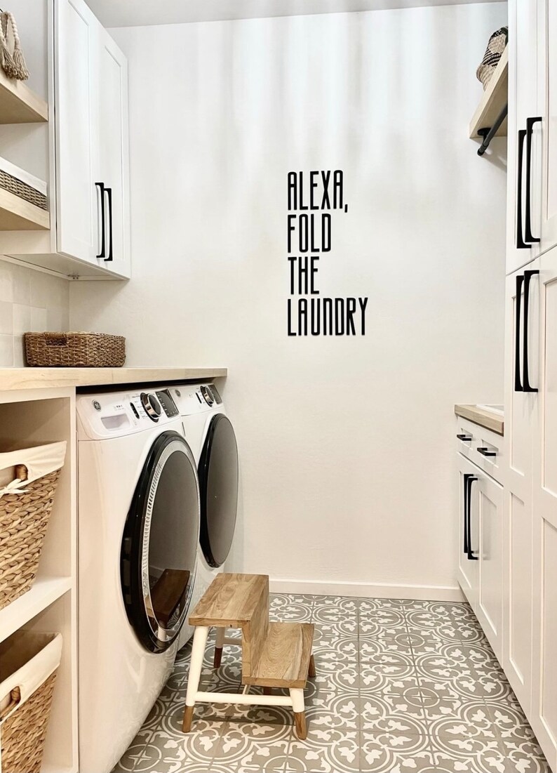 Puede incluir: Un lavadero blanco con una lavadora y una secadora, un taburete de madera y un cartel que dice "Alexa, dobla la ropa".