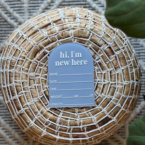 May include: A light blue baby announcement sign with the text "hi, I'm new here" in white. The sign includes spaces to write the baby's name, date, time, weight in pounds and ounces, and length. The sign is placed on a woven basket.