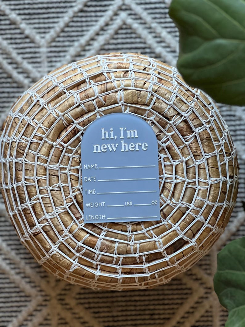 May include: A light blue baby announcement sign with the text "hi, I'm new here" in white. The sign includes spaces to write the baby's name, date, time, weight in pounds and ounces, and length. The sign is placed on a woven basket.
