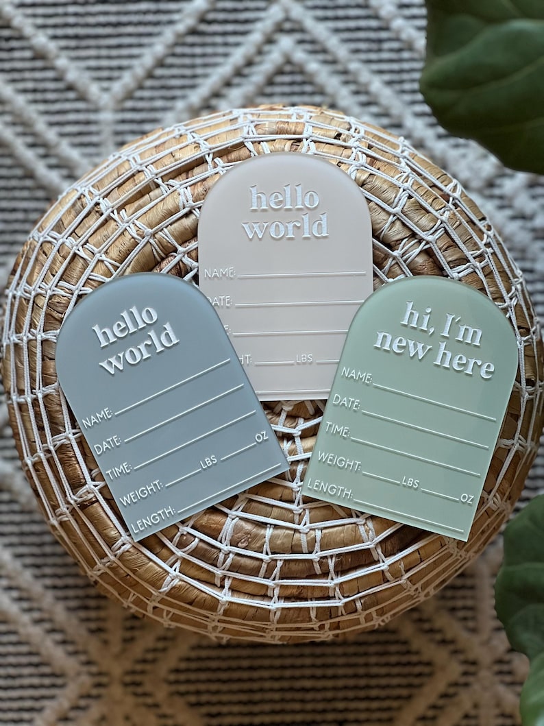 May include: Three pastel-colored baby announcement plaques with rounded tops. The plaques are in shades of gray, beige, and green. Each plaque has the words "hello world" or "hi, I'm new here" in white, along with lines for name, date, time, weight, and length.