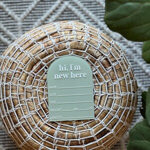 May include: A light green baby announcement tag with the text "hi, I'm new here" in white. The tag includes spaces for name, date, time, weight (lbs, oz), and length. The tag is placed on a woven basket.