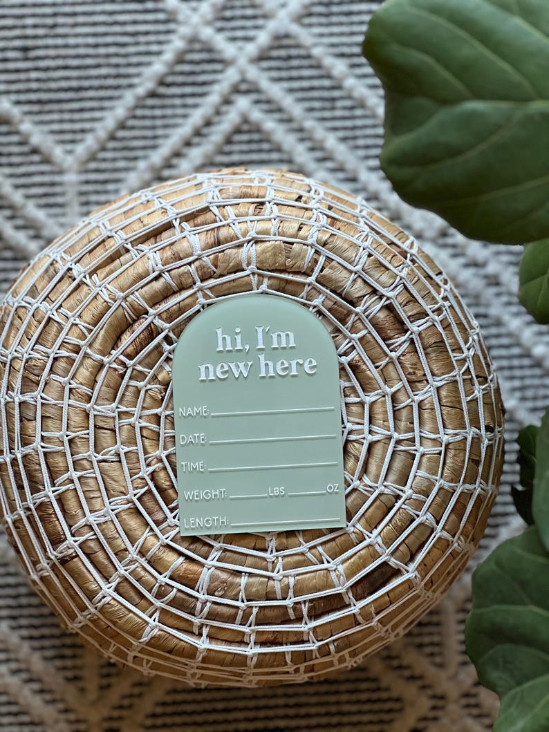 May include: A light green baby announcement tag with the text "hi, I'm new here" in white. The tag includes spaces for name, date, time, weight (lbs, oz), and length. The tag is placed on a woven basket.