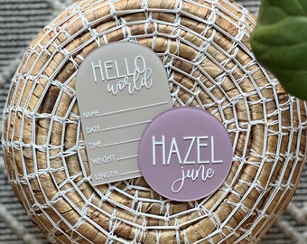 May include: A beige acrylic baby announcement tag with the text "Hello World" and spaces for name, date, time, weight, and length. A purple acrylic tag with the text "Hazel June" is next to it.