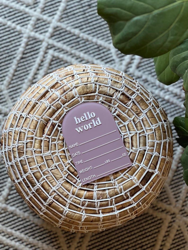 May include: A mauve-colored birth announcement tag with the words "hello world" in white. The tag includes spaces to write the baby's name, date, time, weight (lbs), and length (oz). The tag is on a woven basket.