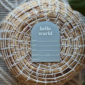 May include: A light blue acrylic birth announcement sign with the words "hello world" in white. The sign has space to write the baby's name, date, time, weight in pounds and ounces, and length. The sign is placed on a woven basket.