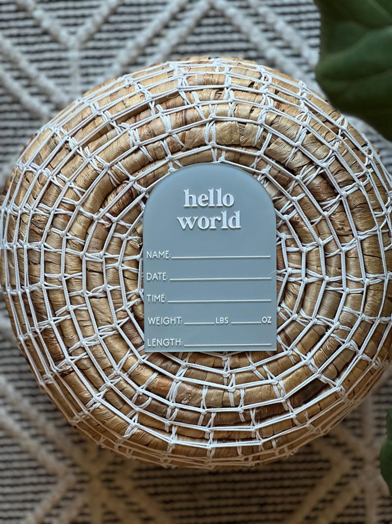 May include: A light blue acrylic birth announcement sign with the words "hello world" in white. The sign has space to write the baby's name, date, time, weight in pounds and ounces, and length. The sign is placed on a woven basket.