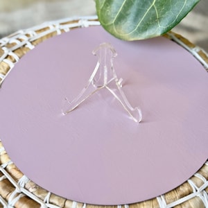May include: A clear acrylic display stand is centered on a round, light purple surface. The surface sits atop a woven basket with a white rope trim. A large green leaf is in the background.