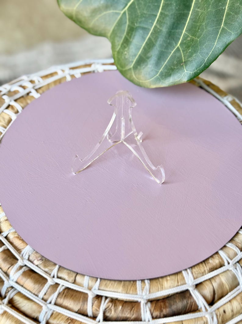 May include: A clear acrylic display stand is centered on a round, light purple surface. The surface sits atop a woven basket with a white rope trim. A large green leaf is in the background.
