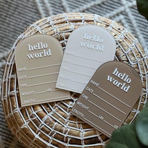 May include: Three acrylic baby announcement plaques in beige, white, and brown, each with the words "hello world" in white. The plaques have space for name, date, time, weight (lbs), and length (inches) details. They are arranged on a woven basket.