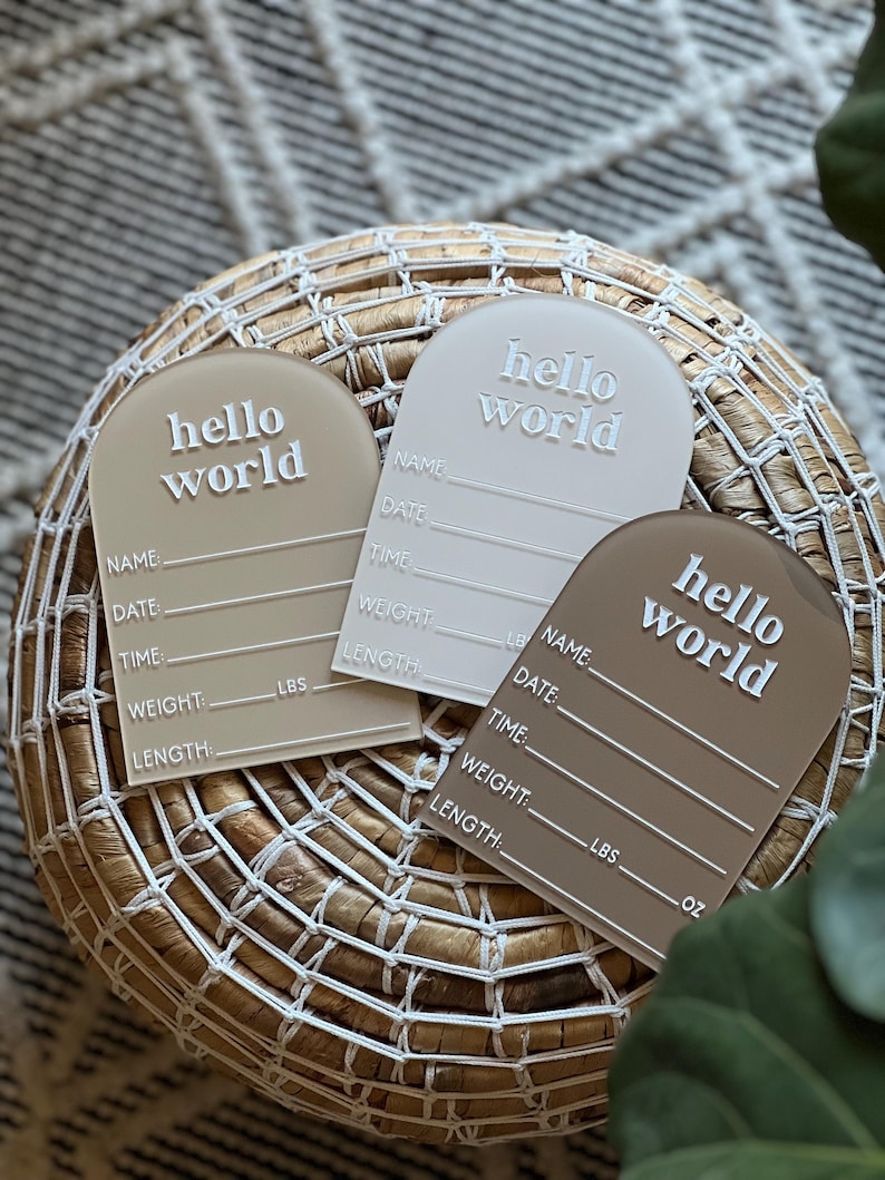 May include: Three acrylic baby announcement plaques in beige, white, and brown, each with the words "hello world" in white. The plaques have space for name, date, time, weight (lbs), and length (inches) details. They are arranged on a woven basket.