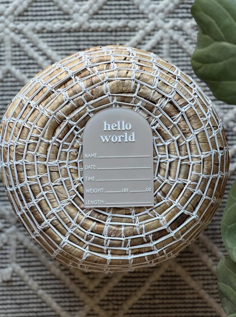 May include: A gray baby announcement plaque with the words "hello world" in white script. The plaque has spaces for name, date, time, weight (lbs, oz), and length. The plaque is set on a woven basket.