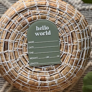 May include: An olive green birth announcement plaque with the words "hello world" in white script. The plaque has lines for name, date, time, weight (lbs, oz), and length. The plaque is placed on a woven basket.