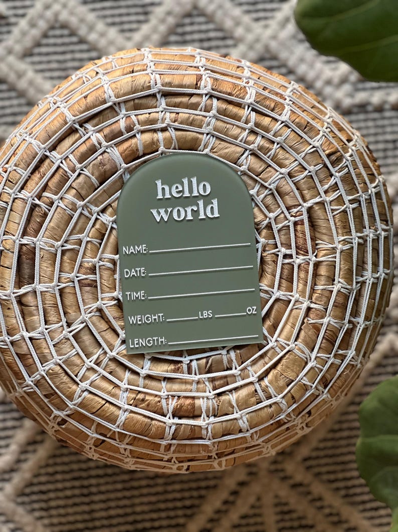 May include: An olive green birth announcement plaque with the words "hello world" in white script. The plaque has lines for name, date, time, weight (lbs, oz), and length. The plaque is placed on a woven basket.