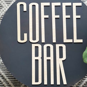 Puede incluir: Un letrero redondo de color gris oscuro con las palabras "COFFEE BAR" en letras de madera blancas.