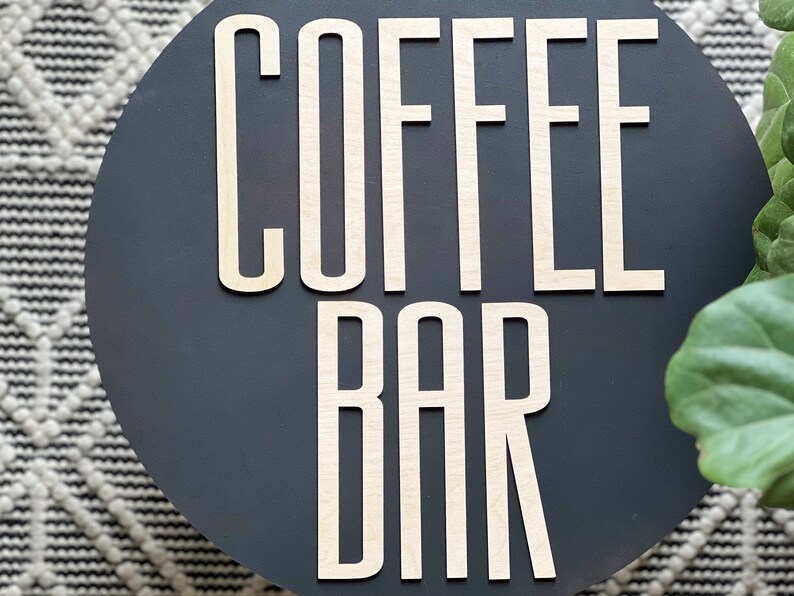 Puede incluir: Un letrero redondo de color gris oscuro con las palabras "COFFEE BAR" en letras de madera blancas.