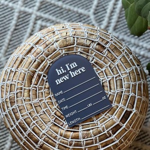 May include: A navy blue birth announcement tag with the text "hi, I'm new here" in white. The tag includes spaces to write the baby's name, date, time, weight in pounds and ounces, and length.