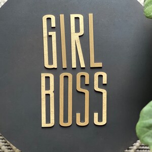 Puede incluir: Un letrero redondo negro con las palabras "GIRL BOSS" en letras doradas.