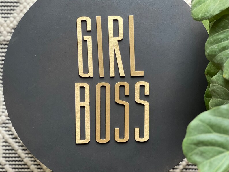 Puede incluir: Un letrero redondo negro con las palabras "GIRL BOSS" en letras doradas.
