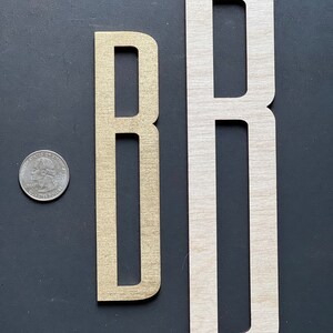 Puede incluir: Dos letras de madera, ambas la letra "B", una es una versi&oacute;n m&aacute;s peque&ntilde;a de color dorado y la otra es una versi&oacute;n m&aacute;s grande de madera natural.