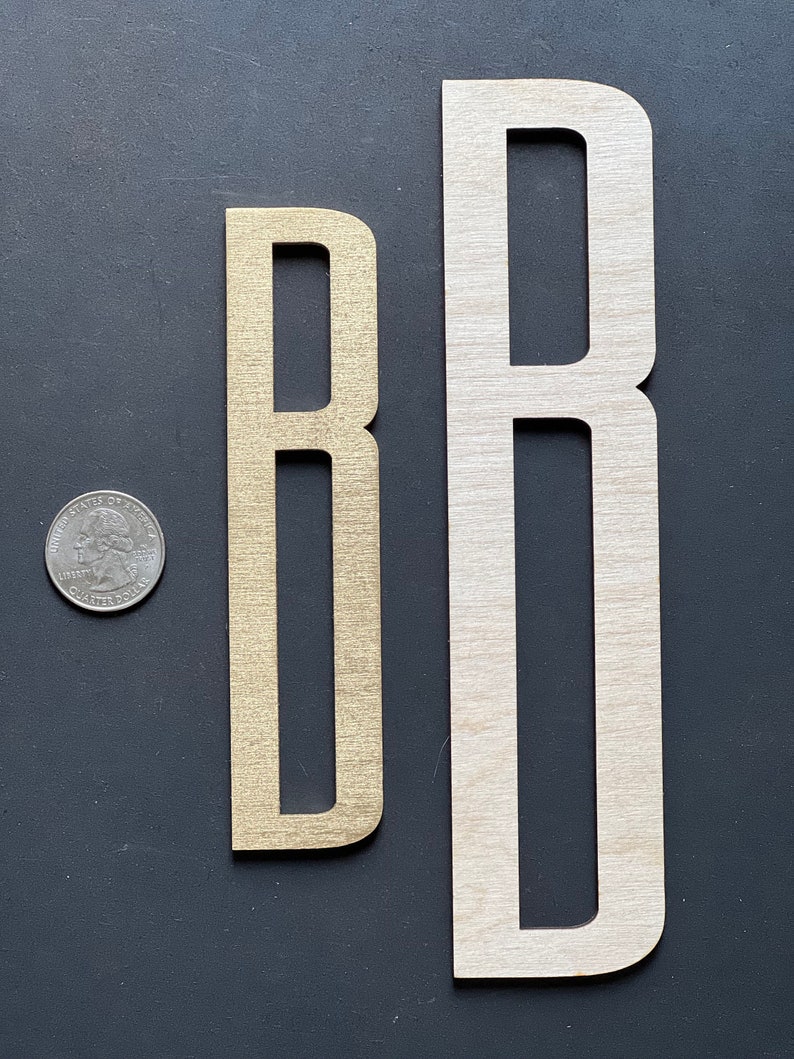 Puede incluir: Dos letras de madera, ambas la letra "B", una es una versi&oacute;n m&aacute;s peque&ntilde;a de color dorado y la otra es una versi&oacute;n m&aacute;s grande de madera natural.
