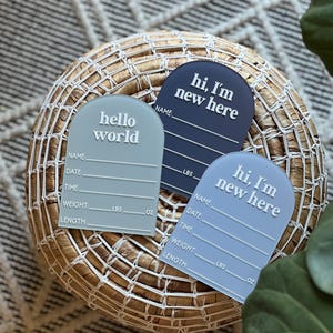 May include: Three pastel-colored baby announcement plaques with the phrases "hello world" and "hi, I'm new here" in white. Each plaque includes spaces for name, date, time, weight (lbs, oz), and length. The plaques are arranged on a woven basket.
