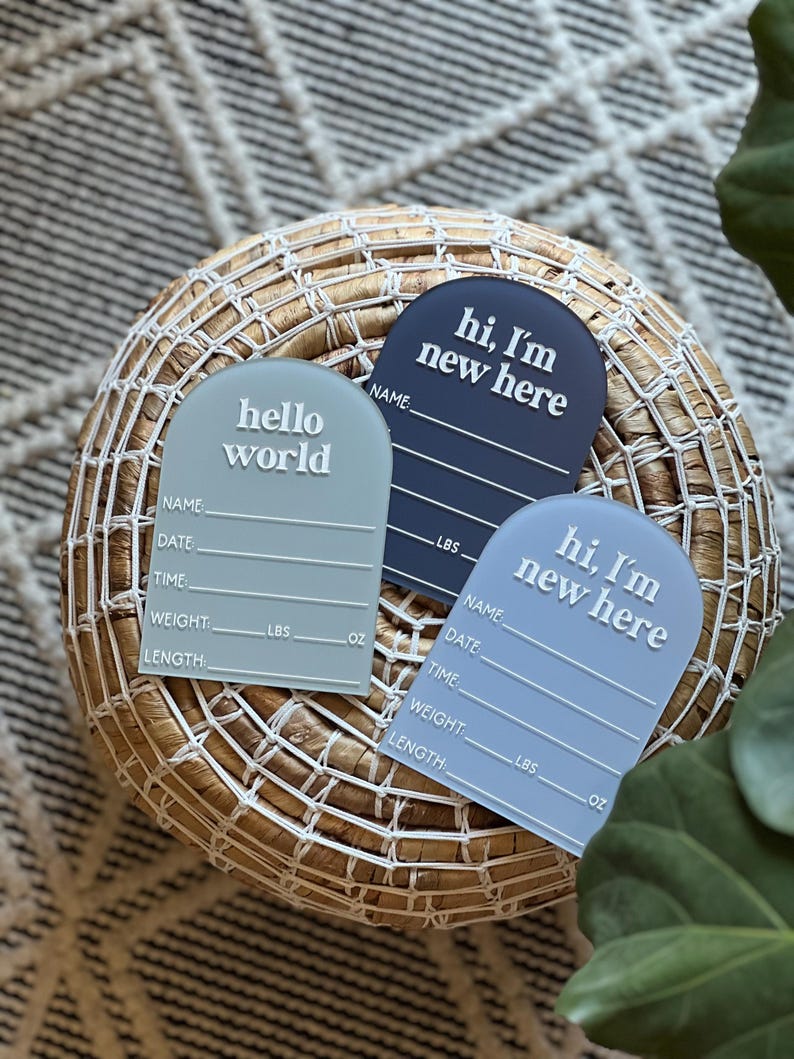 May include: Three pastel-colored baby announcement plaques with the phrases "hello world" and "hi, I'm new here" in white. Each plaque includes spaces for name, date, time, weight (lbs, oz), and length. The plaques are arranged on a woven basket.