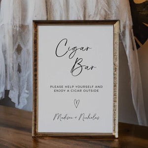 Puede incluir: Un cartel blanco con texto negro que dice "Cigar Bar" y "Por favor, sírvete y disfruta de un cigarro afuera." También se incluye un símbolo de corazón y los nombres "Madison + Nicholas" en el cartel.
