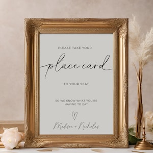 May include: A gold-framed sign with a white background and black text that reads "PLEASE TAKE YOUR place card TO YOUR SEAT SO WE KNOW WHAT YOU'RE HAVING TO EAT ♡ Madison + Nicholas".