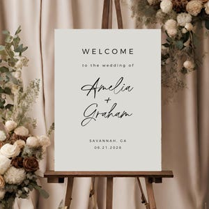 May include: A white welcome sign with black text that reads "Welcome to the wedding of Amelia + Graham Savannah, GA 06.21.2028".