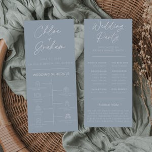 May include: Two light blue wedding invitation cards with white text. The left card has the couple's names, "Chloe + Graham", the wedding date, "June 21, 2028", the location, "La Jolla Beach, California", and a wedding schedule with icons. The right card has the title "Wedding Party", the officiant's name, "Father Daniel Smith", and a list of the wedding party members with their names.