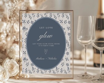 May include: A framed sign with the words "LET LOVE glow" in elegant script. The sign has a blue oval with white text and a floral background. Two wine glasses and a champagne bottle are in the background.