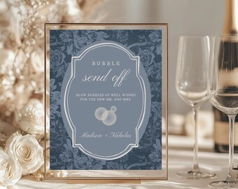 May include: A framed sign with the words "BUBBLE send off" in elegant script. The sign is a soft blue with a floral pattern and includes the names "Madison + Nicholas". The sign is in a gold frame, with champagne flutes and a bottle of champagne in the background.