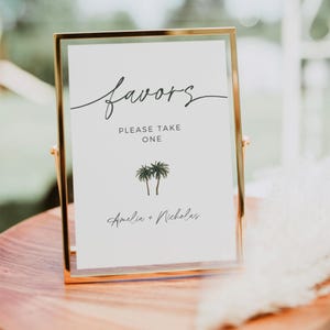 May include: A gold-framed sign with the text "favors" in cursive script. Below the text, it reads "Please take one" and features a small illustration of two palm trees. The sign is finished with the names "Amelia + Nicholas" in cursive script.