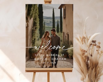 May include: A wedding welcome sign with a photo of a couple in front of a building with a white cursive font that says "welcome to the wedding of Amelia + Graham 08.21.2028"