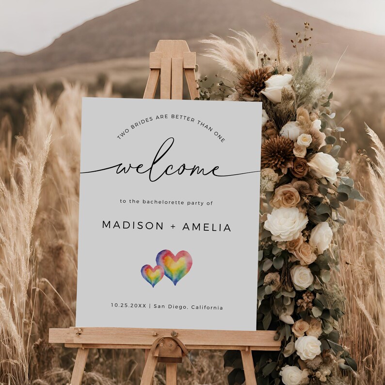 May include: A white welcome sign with the text "Two brides are better than one" and "welcome to the bachelorette party of Madison + Amelia". The sign also includes the date "10.25.20XX" and the location "San Diego, California". The sign is decorated with two rainbow watercolor hearts.
