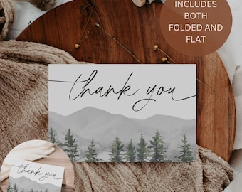 May include: A thank you card with a watercolor illustration of a mountain range and pine trees. The card is on a wooden surface with a brown fabric background. The text "thank you" is written in black cursive script. The text "Includes both folded and flat" is written in brown text on a brown circle.