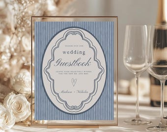 May include: A framed wedding guestbook sign with a blue striped background. The sign features an oval design with the words "wedding Guestbook" and "Madison + Nicholas". The sign is in a gold frame.